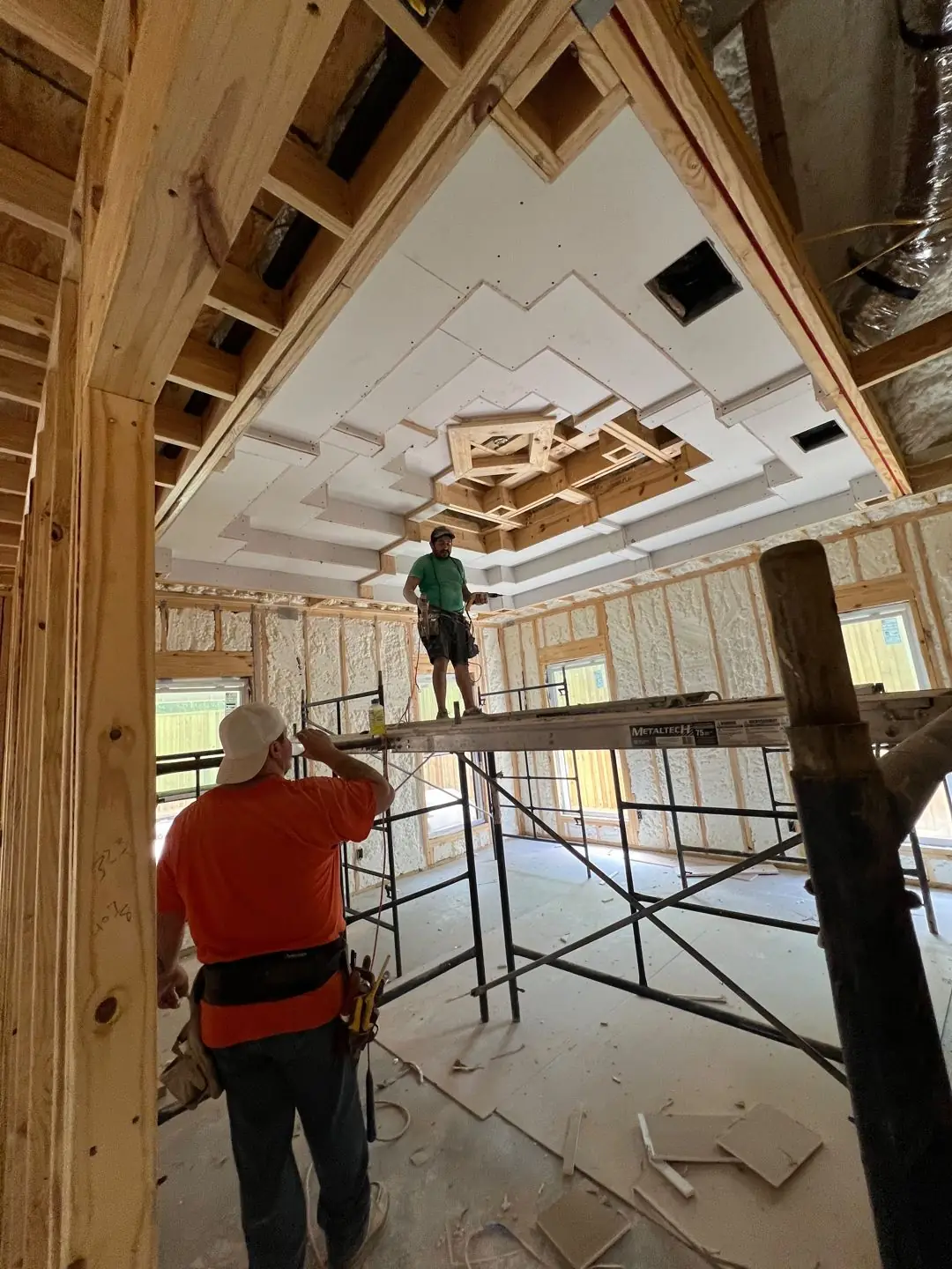 Framing / spray foam / insulation / drywall stages: tray ceilings, scaffold, workers, plumbing PEX manifold, electrical conduit